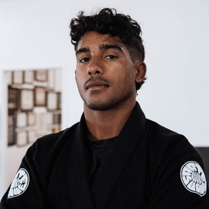 Person in kimono black gi with white sleeve Arte Suave Hooks emblems, standing against a wall