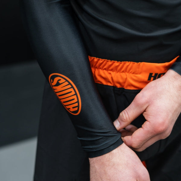 Hooks Long Sleeve Rash Guard Black Inferno, close-up of orange oval sleeve logo, worn showing orange waistband detail