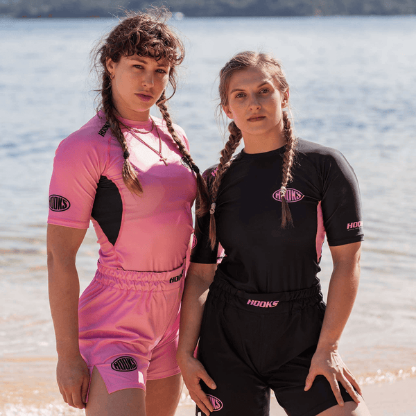 Hooks Short Sleeve Rash Guard Rose Fusion, worn by two people showing pink and black colourways side by side at a beach.