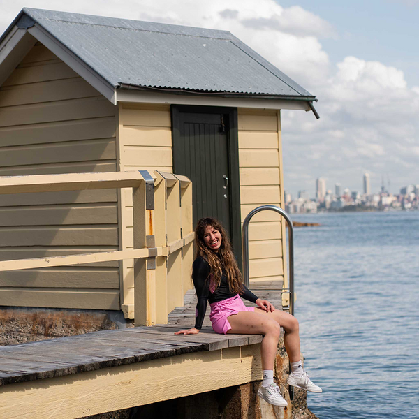 Hooks Long Sleeve Rash Guard Rose Noir, worn by a person seated on a waterfront pier, paired with pink shorts.