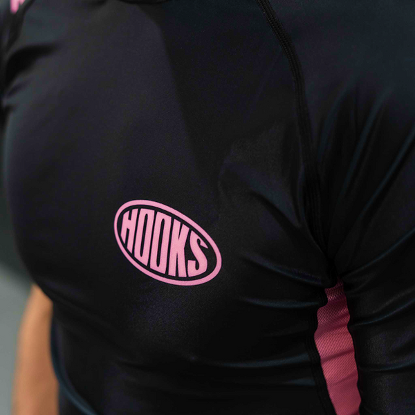 Hooks Short Sleeve Rash Guard Rose Noir, close-up of chest logo — pink oval Hooks badge on black fabric, worn.