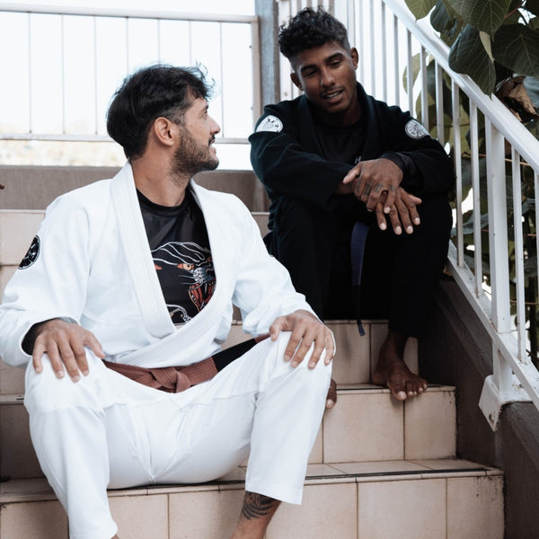 Two martial artists in white and black gis sitting on outdoor stairs, with patches on their arms and a relaxed atmosphere